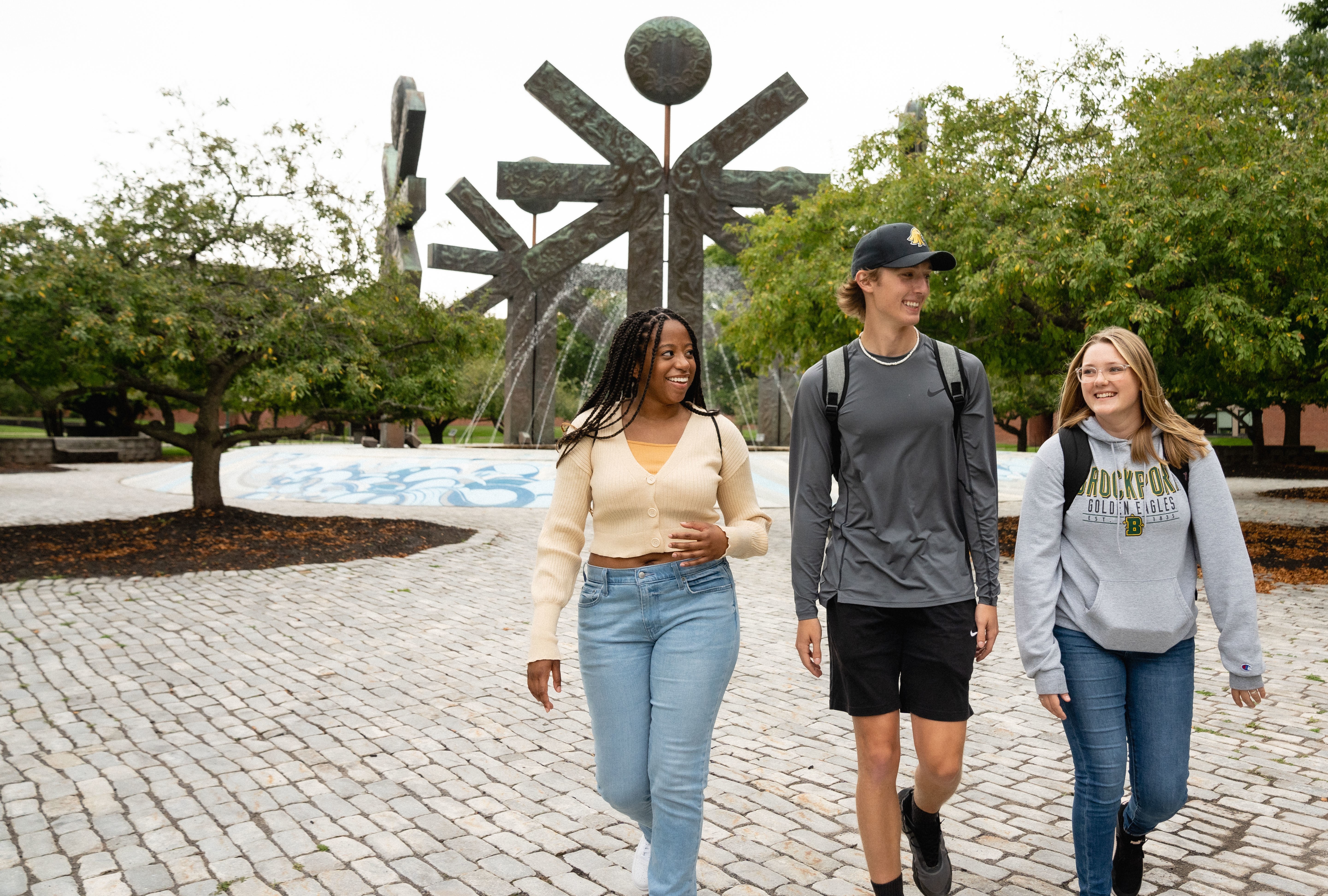 three students smiling and walking in front of fountain with backpacks