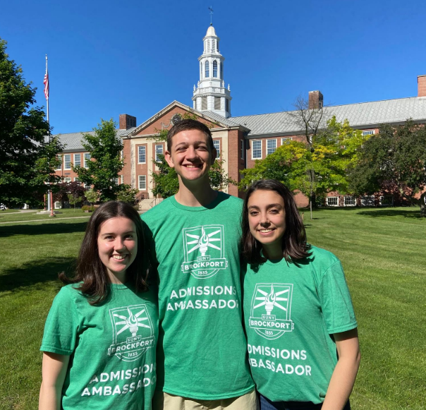 three smiling students in Brockport shirts, with a brick building with a bell tower behind them